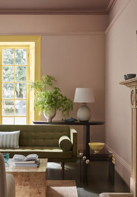 Living space painted in muted pink shade 'Masquerade' with a contrasting yellow 'Light Gold' window frame and an olive green sofa in the foreground.