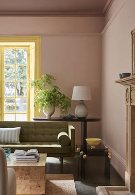 Living space painted in muted pink shade 'Masquerade' with a contrasting yellow 'Light Gold' window frame and an olive green sofa in the foreground.